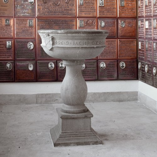 La pila dell'acqua santa della vecchia chiesa, datata 1569; sullo sfondo le targhe ricordo dei caduti