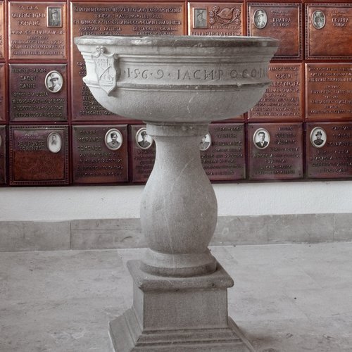 La pila dell'acqua santa della vecchia chiesa, datata 1569; sullo sfondo le targhe ricordo dei caduti